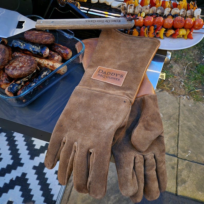 K&ouml;nnte beinhalten: Ein Paar braune Leder-Grillhandschuhe mit dem Text "Daddy's BBQ Gloves" auf der R&uuml;ckseite eines Handschuhs.