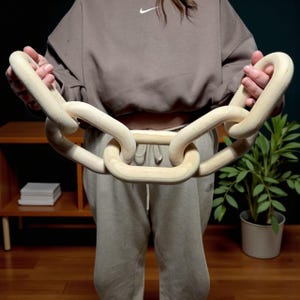 Op de afbeelding: Een grote, decoratieve houten ketting met in elkaar grijpende schakels. De ketting is gemaakt van lichtgekleurd hout en heeft een gladde, natuurlijke afwerking. De schakels zijn afgerond en substantieel, waardoor een gedurfde, moderne esthetiek ontstaat. De ketting wordt voor een persoon gehouden.