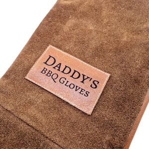 K&ouml;nnte beinhalten: Braune Leder-Grillhandschuhe mit einem gestickten Etikett mit der Aufschrift "Daddy's BBQ Gloves".