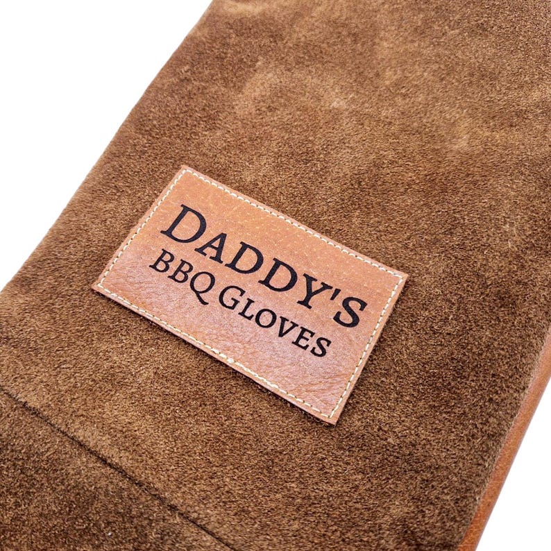 K&ouml;nnte beinhalten: Braune Leder-Grillhandschuhe mit einem gestickten Etikett mit der Aufschrift "Daddy's BBQ Gloves".