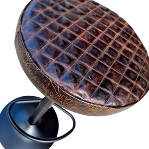 May include: A brown, diamond-patterned leather seat stool with a black metal base. The seat has a distressed appearance, and the stool has a footrest. The design is reminiscent of a vintage or industrial style.