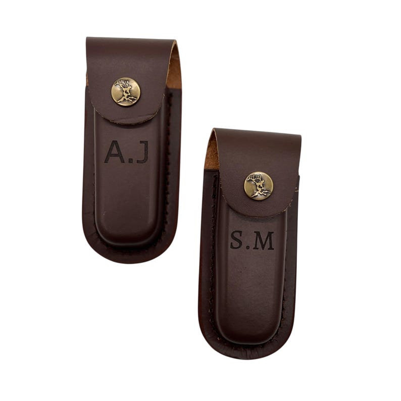 May include: Two brown leather knife sheaths with brass-colored rivets and a tree emblem. One sheath has the initials "A.J" and the other has "S.M" engraved on the front. The sheaths are closed with a flap.
