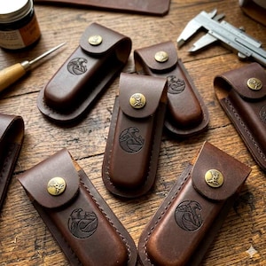 May include: A collection of brown leather knife sheaths, each featuring a wave and mountain motif, displayed on a wooden surface. The sheaths have brass button closures. Crafting tools and a dye jar are also visible.