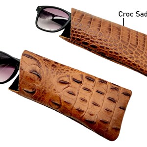 May include: Two brown leather sunglass cases with a crocodile pattern. The case on the top has the text "Croc Saddle" written on the right side of the image.
