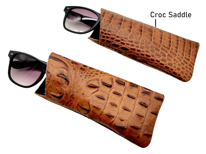 May include: Two brown leather sunglass cases with a crocodile pattern. The case on the top has the text "Croc Saddle" written on the right side of the image.