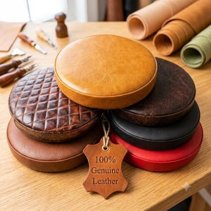 May include: A stack of round leather cushions in various colors including tan, brown, black, and red, with one featuring a quilted pattern. A tan leather tag hangs from the stack, embossed with the text 100% Genuine Leather in black ink.