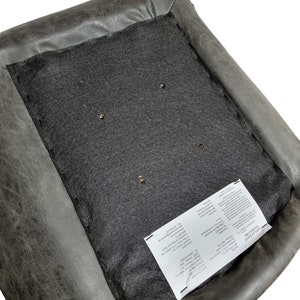 K&ouml;nnte beinhalten: Nahaufnahme eines grauen Lederstuhlsitzes mit freiliegendem Innenraum. Die Unterseite zeigt einen dunkelgrauen Stoff und mehrere kleine L&ouml;cher. Ein wei&szlig;es Etikett mit Text ist am Stoff befestigt.