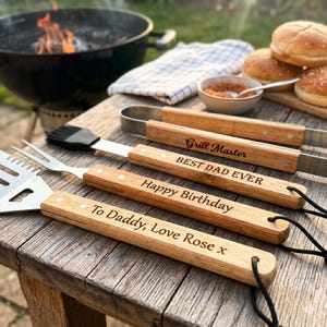 Può includere: Set di utensili da barbecue con manici in legno e testo inciso, tra cui una spatola, un pennello e delle pinze. I manici sono marrone chiaro e riportano scritte come "Grill Master" e "Happy Birthday". Un barbecue e panini sullo sfondo.