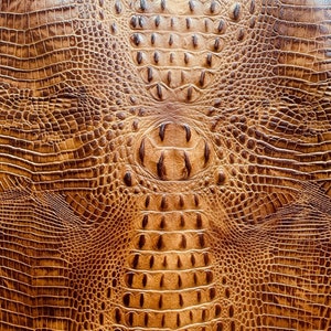 May include: Close-up of a textured, brown leather material with a reptile-like pattern. The design features a repeating scale-like texture, with a central, rounded area and a darker, textured pattern.