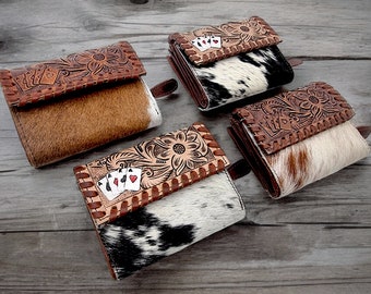 May include: Four leather wallets with cowhide accents. Each wallet features a brown, tooled leather flap with floral designs and playing card details. The wallets have different cowhide patterns in shades of brown, black, and white.