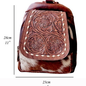 May include: A brown leather backpack with a cowhide flap. The flap has intricate floral tooling and is accented with white stitching. The backpack measures 11 inches tall and 9 inches wide.