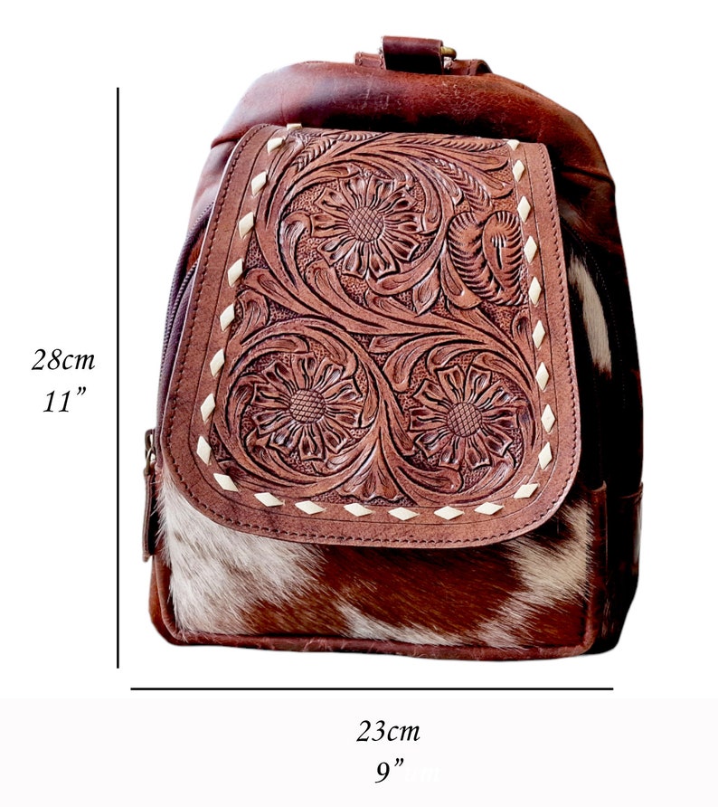 May include: A brown leather backpack with a cowhide flap. The flap has intricate floral tooling and is accented with white stitching. The backpack measures 11 inches tall and 9 inches wide.