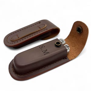 May include: A brown leather multi-tool case with a matching belt clip. The case is open, revealing a silver multi-tool. The initials "S.M" are embossed on the case. The case is made of brown leather with a stitched border.