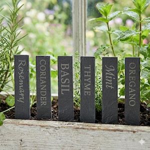 May include: Six rectangular slate herb garden markers with engraved white lettering. The markers read: Rosemary, Coriander, Basil, Thyme, Mint, and Oregano. The markers are placed in a wooden planter box with green herbs and plants.