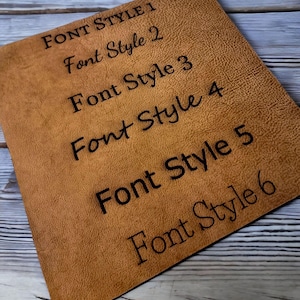 May include: A square, tan leather swatch with six different font styles printed in black. The text reads "FONT STYLE 1" through "FONT STYLE 6". The leather has a textured surface.