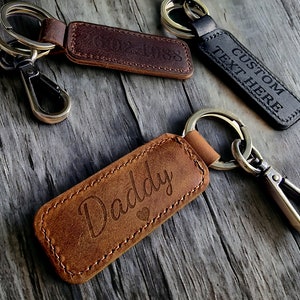 Può includere: Tre portachiavi in ​​pelle con anelli e chiusure in metallo. I portachiavi sono marroni e neri e hanno diversi testi incisi: "Daddy", "Testo personalizzato qui" e "02-1988".
