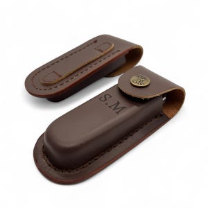 May include: Two brown leather knife sheaths. One is open, displaying the interior, and the other is closed. The open sheath has a loop for a belt. The closed sheath has a brass button closure with a tree design and the initials "S.M."