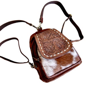 May include: A brown leather backpack with a white and brown cowhide flap. The flap has a floral design embossed into the leather. The backpack has a long adjustable strap.