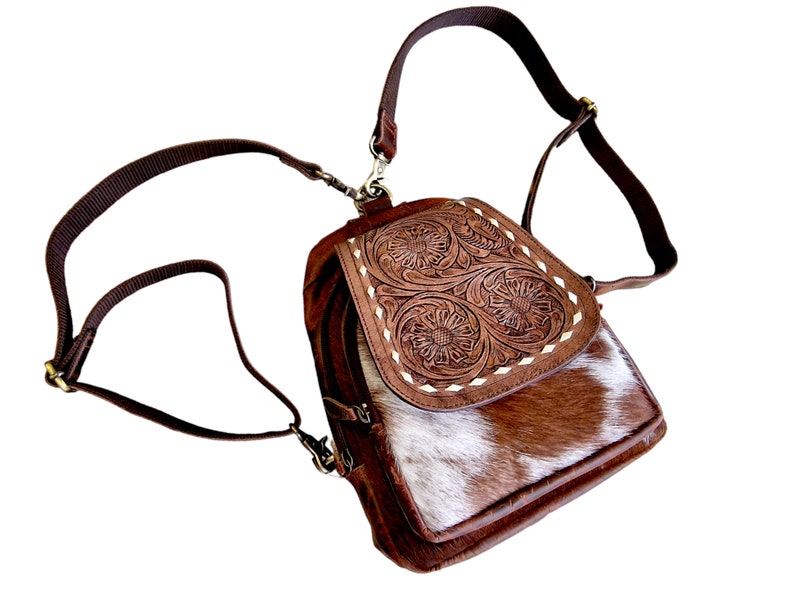 May include: A brown leather backpack with a white and brown cowhide flap. The flap has a floral design embossed into the leather. The backpack has a long adjustable strap.