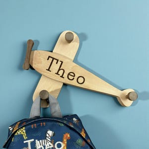 May include: Wooden airplane-shaped wall hook with the name "Theo" engraved on the body. The hook has a propeller and three wooden pegs for hanging items. A blue backpack with a jungle animal pattern hangs from the hook.