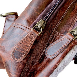 May include: Close-up of a brown leather backpack with two zippers and a flap. The zippers have metal pulls and the flap has a stitched detail.