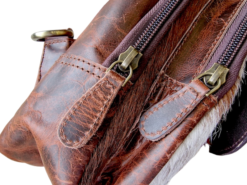 May include: Close-up of a brown leather backpack with two zippers and a flap. The zippers have metal pulls and the flap has a stitched detail.