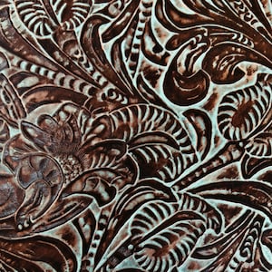 Op de afbeelding: Close-up van een bruine lederen textuur met een gegraveerd bloemenpatroon. Het leer heeft een lichtblauwe achtergrond en het patroon is reliëf en gedetailleerd.