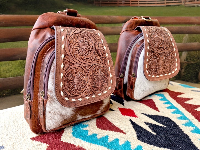 May include: Two brown leather backpacks with intricate floral tooled designs. The backpacks have a white cowhide patch on the front and are sitting on a colorful rug.