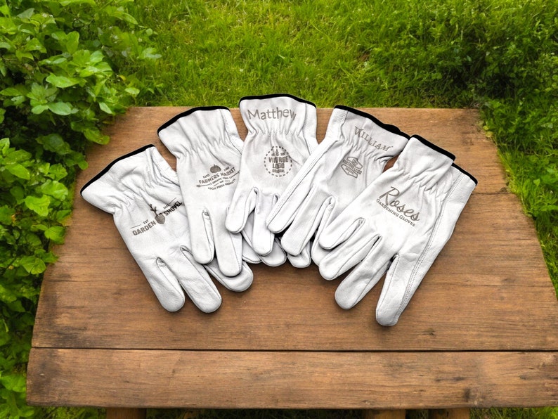 May include: Five pairs of white leather gardening gloves with personalized names and logos. The gloves have black trim and are laid out on a wooden surface.