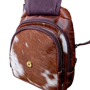 May include: A brown leather backpack with a white and brown cowhide pattern. The backpack has a zippered closure and a single strap.