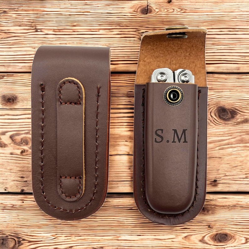 May include: Two brown leather multi-tool cases. One case is closed, featuring a belt loop on the back. The other case is open, revealing a silver multi-tool. The initials "S.M" are embossed on the front of the open case.
