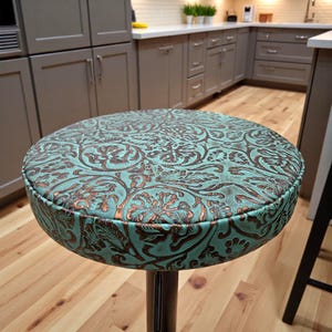 May include: A round bar stool with a turquoise and brown patterned seat. The seat has a detailed floral design and a brown border. The stool has a black metal base and is set in a kitchen with wooden floors.