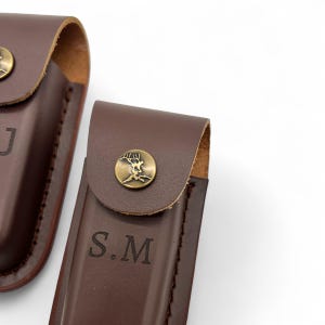 May include: Close-up of two brown leather sheaths. Each sheath has a flap secured by a brass button with a tree design. One sheath is embossed with the initials "S.M."
