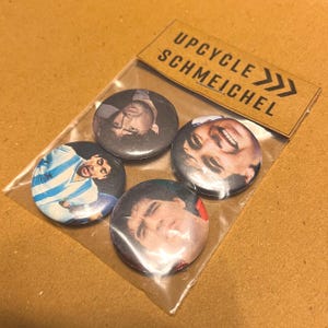 May include: Four circular pin badges featuring images of faces, presented in a clear plastic packet. The badges display various images of people. A brown paper tag above the badges reads "UPCYCLE SCHMEICHEL".