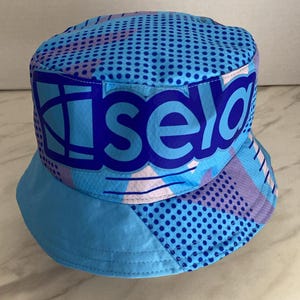 May include: A bucket hat featuring a vibrant blue, light blue, and purple geometric pattern with a prominent dark blue graphic print on the front that reads "isela" in bold, lowercase letters, accented by a repeating dark blue polka dot design.