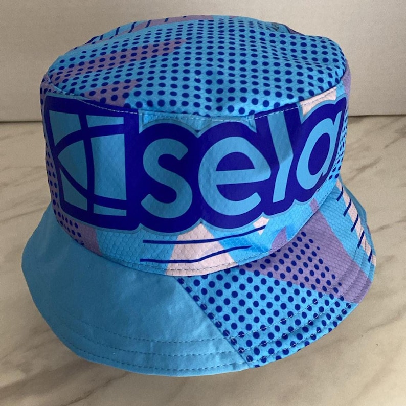 May include: A bucket hat featuring a vibrant blue, light blue, and purple geometric pattern with a prominent dark blue graphic print on the front that reads "isela" in bold, lowercase letters, accented by a repeating dark blue polka dot design.