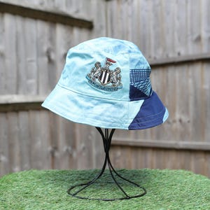 May include: Light blue bucket hat with a Newcastle United crest. The hat features a patchwork design with navy blue and patterned sections. The hat is displayed on a black metal stand against a wooden fence.