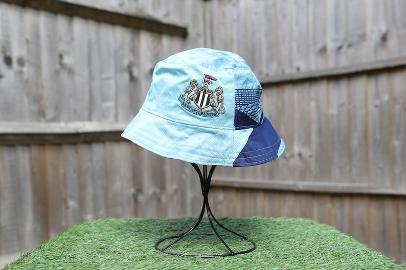 May include: Light blue bucket hat with a Newcastle United crest. The hat features a patchwork design with navy blue and patterned sections. The hat is displayed on a black metal stand against a wooden fence.