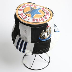 May include: Bucket hat with a black, white, and blue color block design. The hat features a Newcastle Brown Ale logo and a Newcastle United crest. The hat is displayed on a wire stand.
