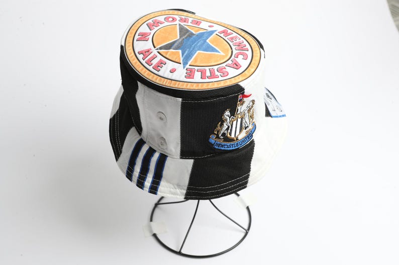 May include: Bucket hat with a black, white, and blue color block design. The hat features a Newcastle Brown Ale logo and a Newcastle United crest. The hat is displayed on a wire stand.