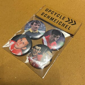 Puede incluir: Un conjunto de cuatro botones redondos con retratos de futbolistas. Los botones están empaquetados en una bolsa de plástico transparente con una etiqueta de cartón marrón que dice "UPCYCLE SCHMEICHEL". Los botones tienen un acabado brillante.