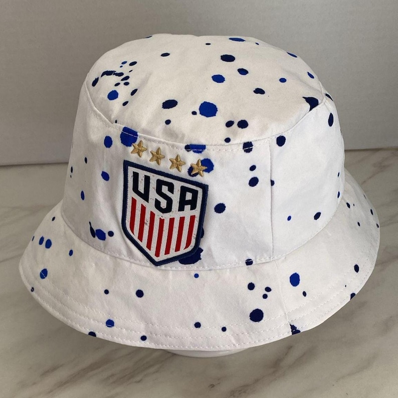 May include: A white bucket hat featuring a blue paint splatter pattern throughout. The front displays a shield-shaped patch with the text USA above red vertical stripes, topped with four embroidered gold stars arranged in a horizontal row.