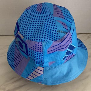 May include: A light blue bucket hat featuring a vibrant geometric pattern in shades of dark blue and purple. The design includes polka dots, diagonal lines, and the text AEROREADY printed on the side. The hat has a wide, downward-sloping brim.