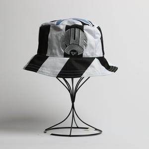 May include: A black and white striped bucket hat with a logo on the front. The hat is displayed on a black wire stand against a white background. The hat has a black brim and a white and black striped pattern.