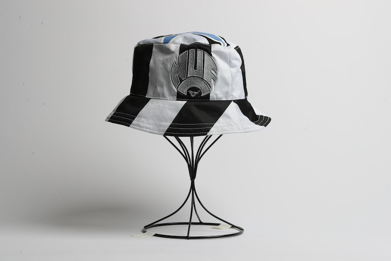 May include: A black and white striped bucket hat with a logo on the front. The hat is displayed on a black wire stand against a white background. The hat has a black brim and a white and black striped pattern.
