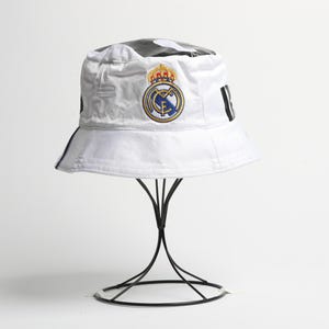 Puede incluir: Sombrero de pescador blanco con el escudo del Real Madrid bordado. El sombrero tiene un patr&oacute;n en blanco y negro en la parte superior y rayas negras en los lados. Se exhibe en un soporte de metal negro.