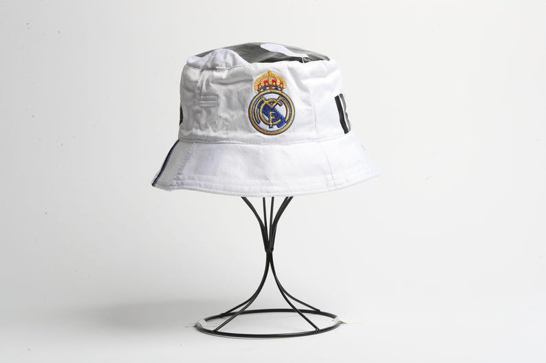 Puede incluir: Sombrero de pescador blanco con el escudo del Real Madrid bordado. El sombrero tiene un patr&oacute;n en blanco y negro en la parte superior y rayas negras en los lados. Se exhibe en un soporte de metal negro.