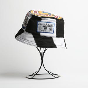 May include: A black and white striped bucket hat with patches and logos, including a Newcastle Football Club badge. The hat is displayed on a black metal stand. The hat has a white interior and a black brim.
