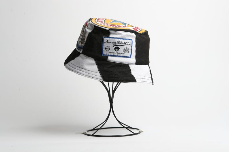 May include: A black and white striped bucket hat with patches and logos, including a Newcastle Football Club badge. The hat is displayed on a black metal stand. The hat has a white interior and a black brim.