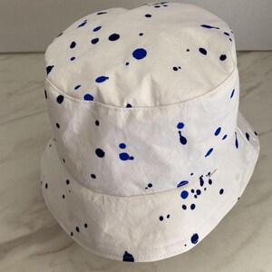May include: A white bucket hat featuring a scattered pattern of dark blue paint splatters across the crown and the downward-sloping brim. The hat has a flat top and a stitched seam detail around the base of the crown, providing a structured shape.